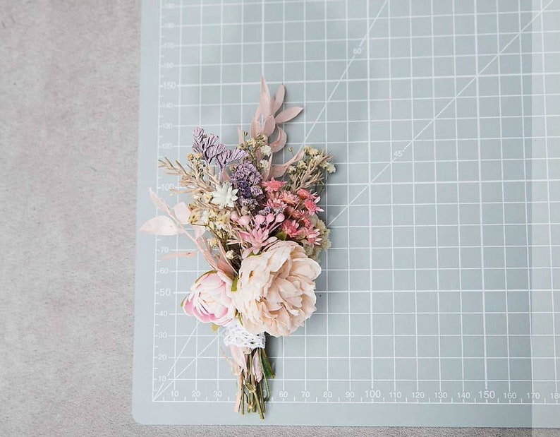May include: A small, dried flower bouquet featuring a variety of colors. The bouquet includes light pink peonies, lavender, and other dried flowers. The stems are wrapped with a white lace ribbon. The bouquet is on a light blue cutting mat.