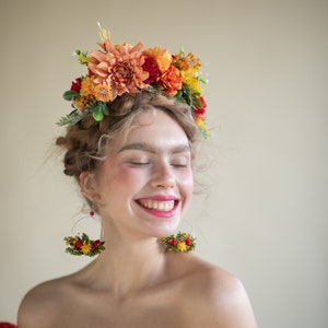 Könnte beinhalten: Eine Frau mit lockigem blonden Haar trägt eine Blumenkrone mit orangefarbenen, roten und gelben Blumen. Sie lächelt und trägt dazu passende florale Creolen. Sie trägt ein rotes Top mit offener Schulter.
