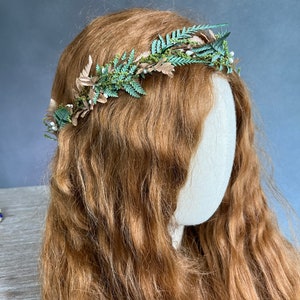 May include: A handcrafted floral crown featuring green fern leaves, tan-colored leaves, and small white berries. The crown is set against a backdrop of long, wavy auburn hair. This headpiece is ideal for weddings, festivals, or special occasions.