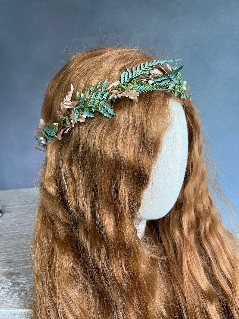 May include: A handcrafted floral crown featuring green fern leaves, tan-colored leaves, and small white berries. The crown is set against a backdrop of long, wavy auburn hair. This headpiece is ideal for weddings, festivals, or special occasions.