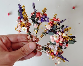Könnte beinhalten: Eine Auswahl an floralen Haarnadeln. Jede Nadel hat einen kleinen Strauß aus getrockneten Blumen in Lila, Gelb, Rot und Weiß. Die Blumen sind an goldfarbenen Haarnadeln befestigt, was einen zarten, natürlichen Look ergibt.