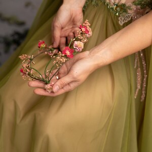 May include: A woman wearing a green dress holds a floral crown made of pink and red flowers.