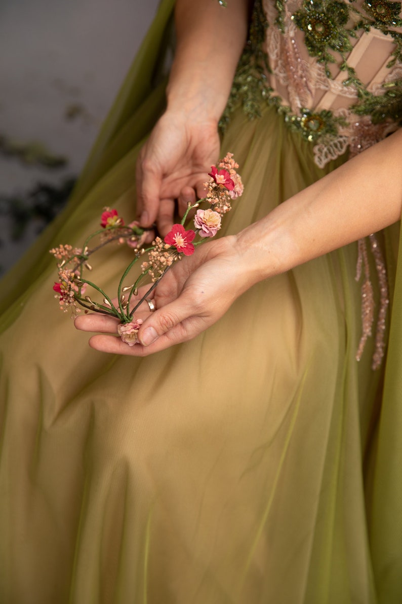 May include: A woman wearing a green dress holds a floral crown made of pink and red flowers.