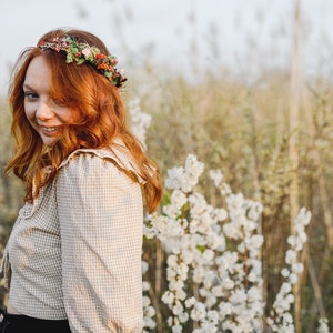 May include: A woman with long, red hair wearing a floral crown and a plaid shirt stands in a field of white flowers.