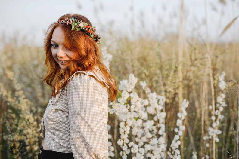 May include: A woman with long, red hair wearing a floral crown and a plaid shirt stands in a field of white flowers.