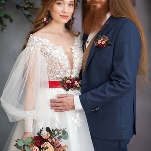 May include: A bride and groom pose for a wedding portrait. The bride is wearing a white lace gown with a red sash and a floral crown. She is holding a bouquet of burgundy and blush flowers. The groom is wearing a navy blue suit with a brown vest and a floral boutonniere.