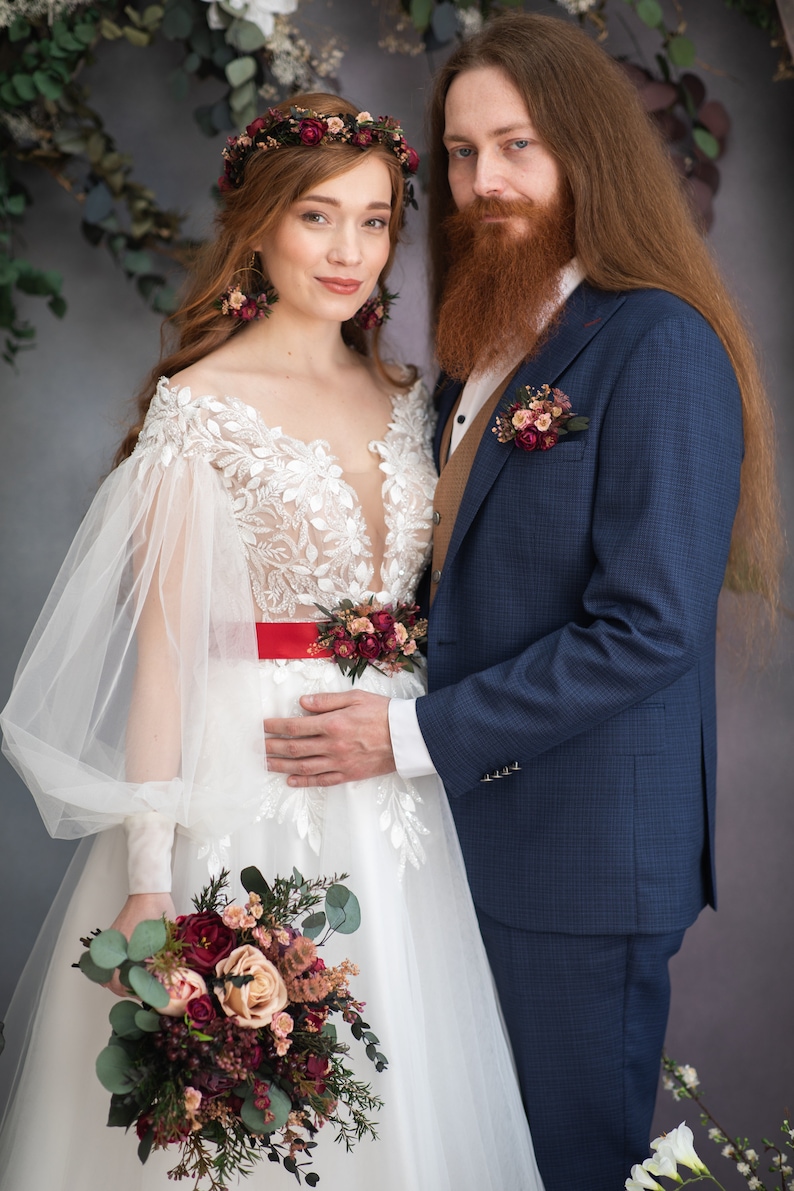 May include: A bride and groom pose for a wedding portrait. The bride is wearing a white lace gown with a red sash and a floral crown. She is holding a bouquet of burgundy and blush flowers. The groom is wearing a navy blue suit with a brown vest and a floral boutonniere.