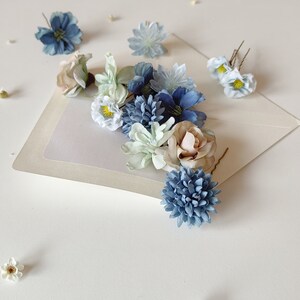K&ouml;nnte beinhalten: Eine Anordnung k&uuml;nstlicher Blumen in Blau-, Wei&szlig;- und Beiget&ouml;nen, auf einer hellen Oberfl&auml;che pr&auml;sentiert. Die Anordnung umfasst verschiedene Blumensorten, wie Rosen und kleine, g&auml;nsebl&uuml;mchenartige Bl&uuml;ten. Einige Blumen sind an Haarnadeln befestigt.