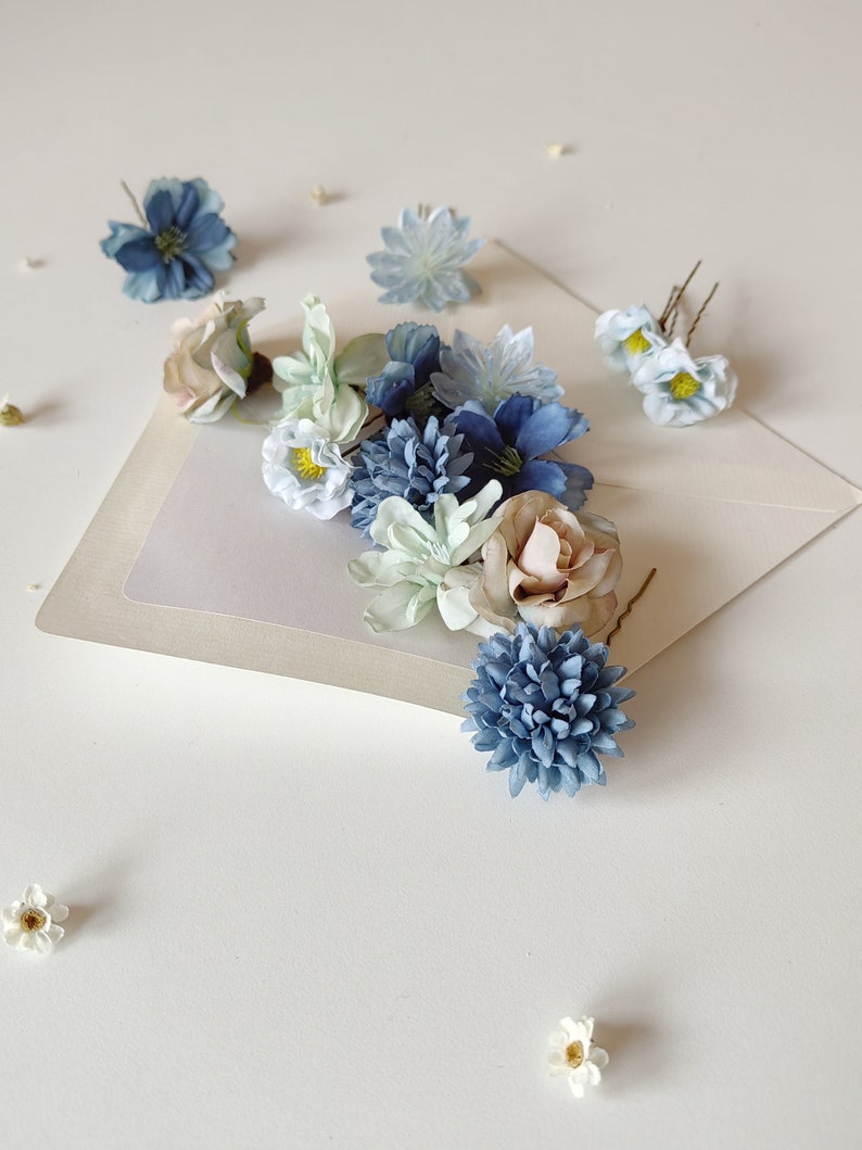 K&ouml;nnte beinhalten: Eine Anordnung k&uuml;nstlicher Blumen in Blau-, Wei&szlig;- und Beiget&ouml;nen, auf einer hellen Oberfl&auml;che pr&auml;sentiert. Die Anordnung umfasst verschiedene Blumensorten, wie Rosen und kleine, g&auml;nsebl&uuml;mchenartige Bl&uuml;ten. Einige Blumen sind an Haarnadeln befestigt.