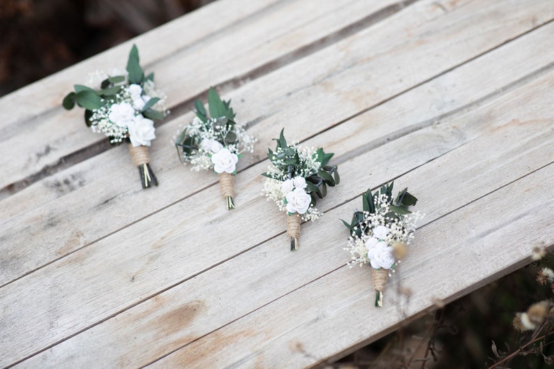 Puede incluir: Cuatro peque&ntilde;os ramos florales con rosas blancas, paniculata y hojas verdes est&aacute;n dispuestos sobre una superficie de madera clara. Los tallos est&aacute;n envueltos con hilo. Los ramos son probablemente para una boda.