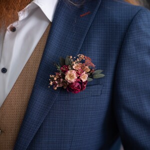 May include: A blue suit jacket with a brown vest and a white shirt. A small bouquet of pink and white flowers is pinned to the lapel.