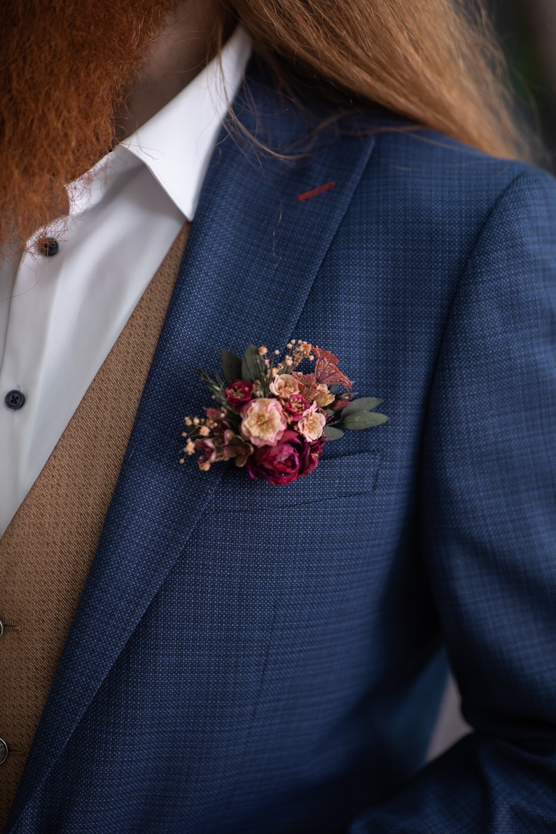 May include: A blue suit jacket with a brown vest and a white shirt. A small bouquet of pink and white flowers is pinned to the lapel.