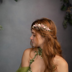 Peut inclure: Une femme aux longs cheveux roux porte une couronne de fleurs avec des fleurs roses et blanches. La couronne est faite de fleurs s&eacute;ch&eacute;es et de verdure. Elle porte une robe verte &agrave; motifs floraux.