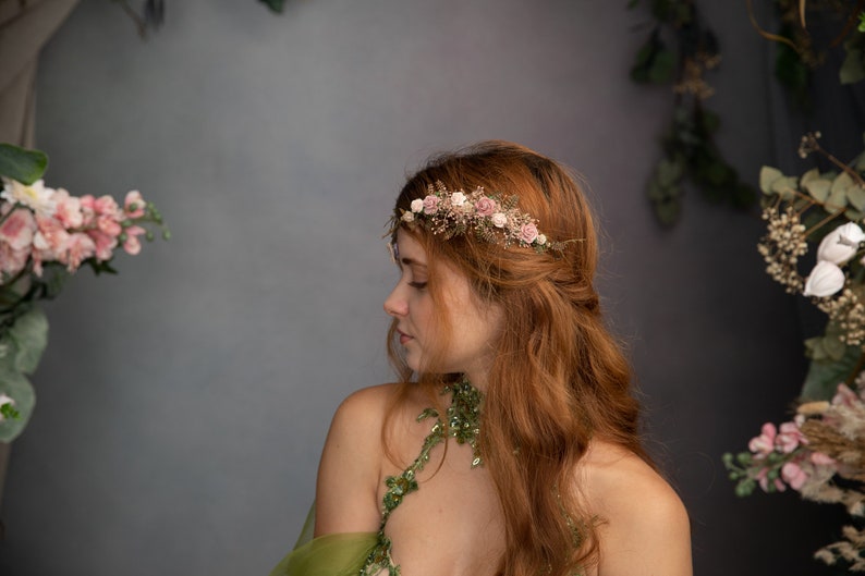 Peut inclure: Une femme aux longs cheveux roux porte une couronne de fleurs avec des fleurs roses et blanches. La couronne est faite de fleurs s&eacute;ch&eacute;es et de verdure. Elle porte une robe verte &agrave; motifs floraux.