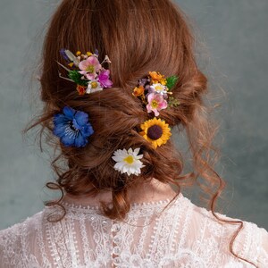 May include: A woman with long, curly red hair wearing a white lace dress with buttons down the back. Her hair is styled in a loose bun with several small flower hair clips.