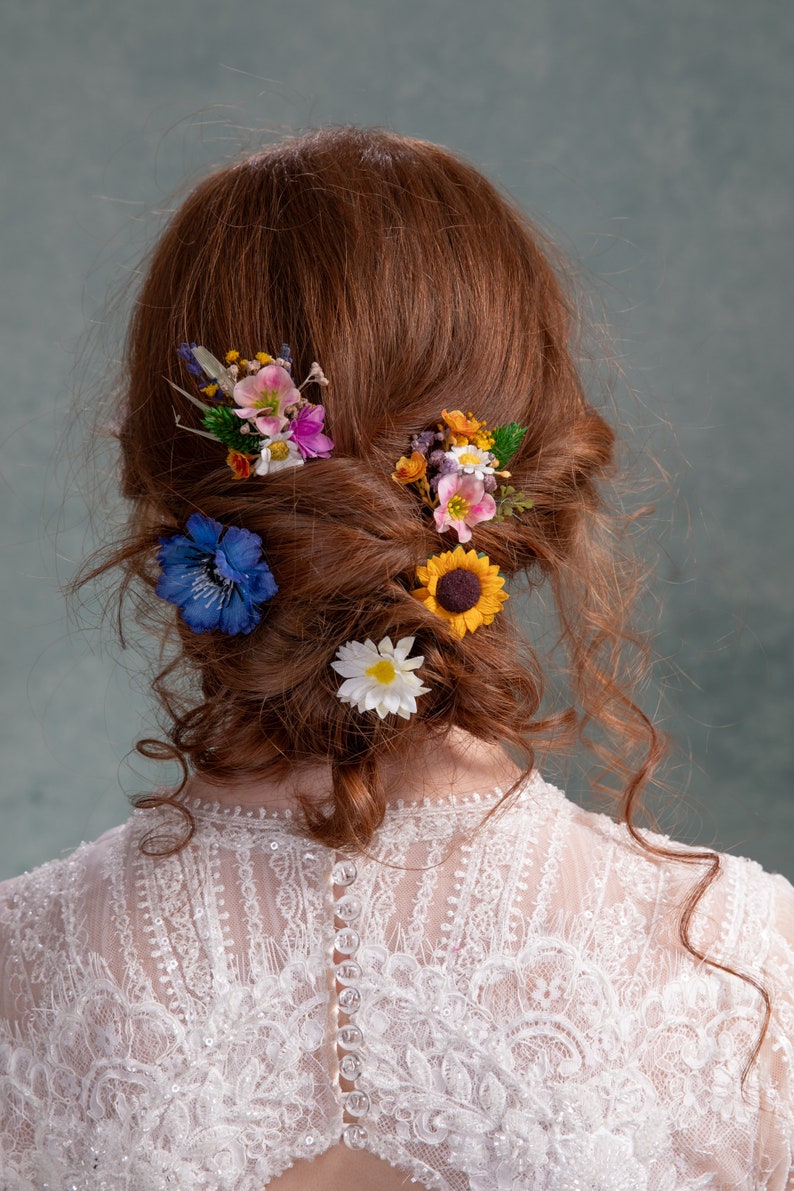 May include: A woman with long, curly red hair wearing a white lace dress with buttons down the back. Her hair is styled in a loose bun with several small flower hair clips.