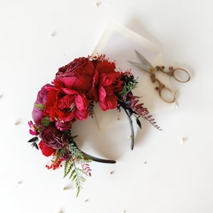 Op de afbeelding: Een bordeauxrode bloemenhoofdband met een zwarte hoofdband. De hoofdband is versierd met bordeauxrode rozen, varens en ander groen.