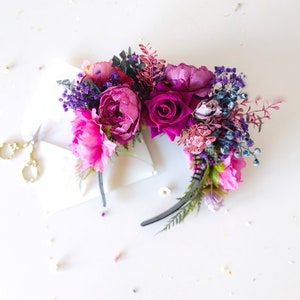 May include: A pink and purple floral headband with a black headband. The headband is decorated with pink roses, purple peonies, and other flowers.