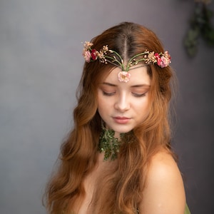 May include: A woman with long, red hair wears a floral crown with pink and red flowers. The crown is made of green vines and has a delicate design. She is wearing a green necklace with a similar floral design.