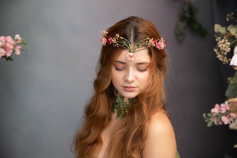 May include: A woman with long, red hair wears a floral crown with pink and red flowers. The crown is made of green vines and has a delicate design. She is wearing a green necklace with a similar floral design.