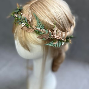 May include: A floral crown featuring brown leaves, green ferns, and small white berries. The crown is set against blonde hair and a white mannequin head. This nature-inspired headpiece is suitable for weddings, festivals, or everyday wear.