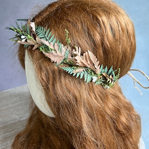 May include: A handmade floral crown featuring a delicate arrangement of faux leaves and greenery. The crown includes light brown, green, and white elements, creating a natural and elegant aesthetic. The crown is secured with twine.