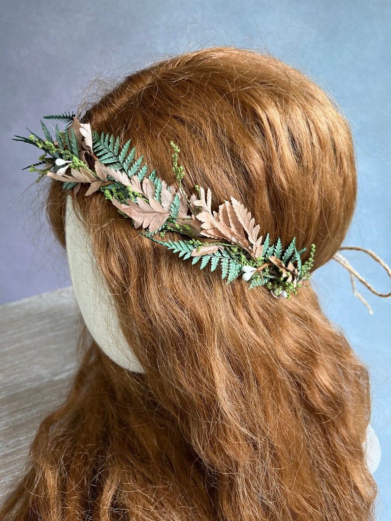 May include: A handmade floral crown featuring a delicate arrangement of faux leaves and greenery. The crown includes light brown, green, and white elements, creating a natural and elegant aesthetic. The crown is secured with twine.