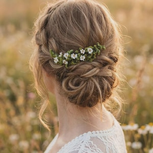 以下が含まれることがあります： 女性のヘアスタイルは、編み込みの王冠と低いお団子で、花のヘアアクセサリーで飾られています。ヘアピースは小さな白い花と緑の葉で構成されています。女性は白いレースのトップスを着ています。