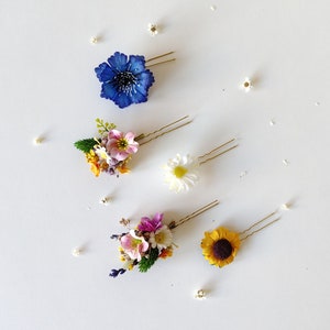 May include: Five floral hair clips with gold bobby pins. The clips feature a variety of flowers, including a blue flower, a pink flower, a white flower, a yellow flower, and a sunflower.