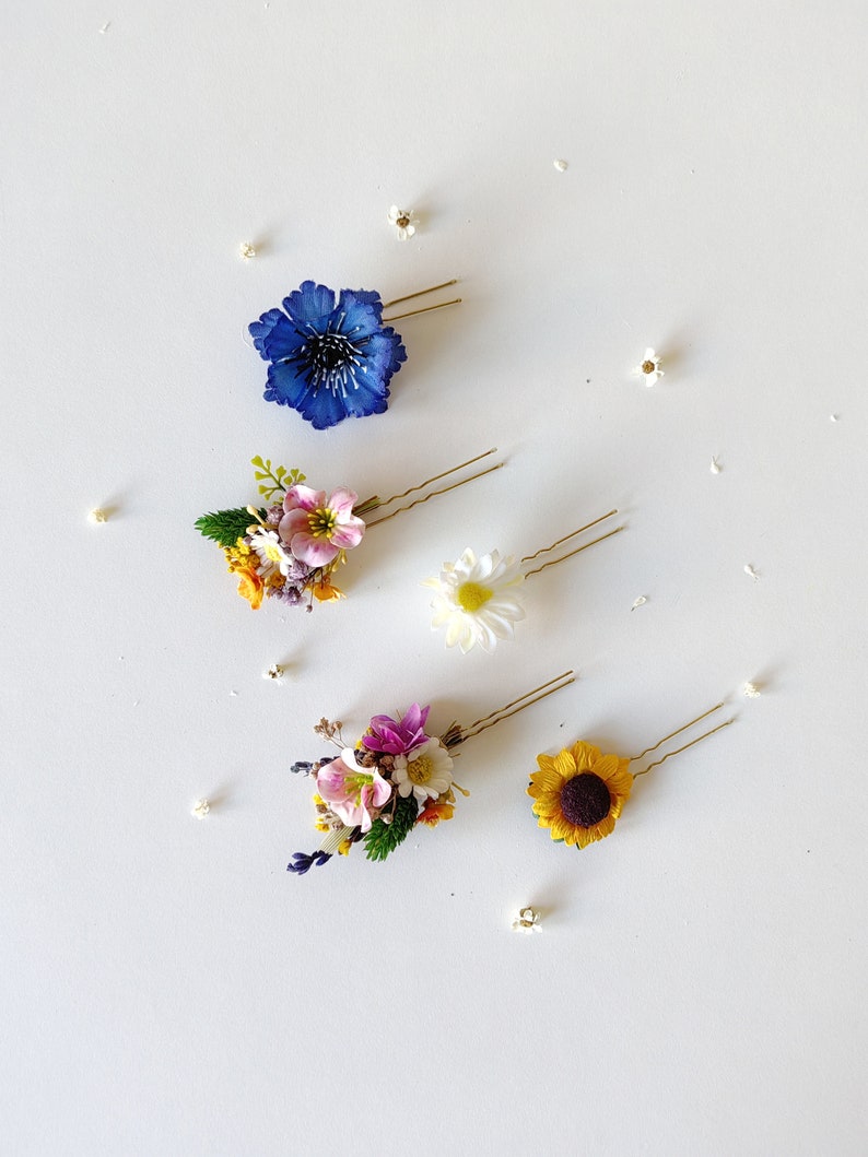 May include: Five floral hair clips with gold bobby pins. The clips feature a variety of flowers, including a blue flower, a pink flower, a white flower, a yellow flower, and a sunflower.
