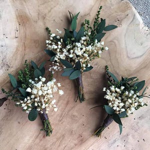 May include: Three small boutonnieres, each with tiny white flowers, green sprigs, and dark green leaves, bound with brown twine. They are displayed on a light brown wooden surface, ideal for weddings or special events.