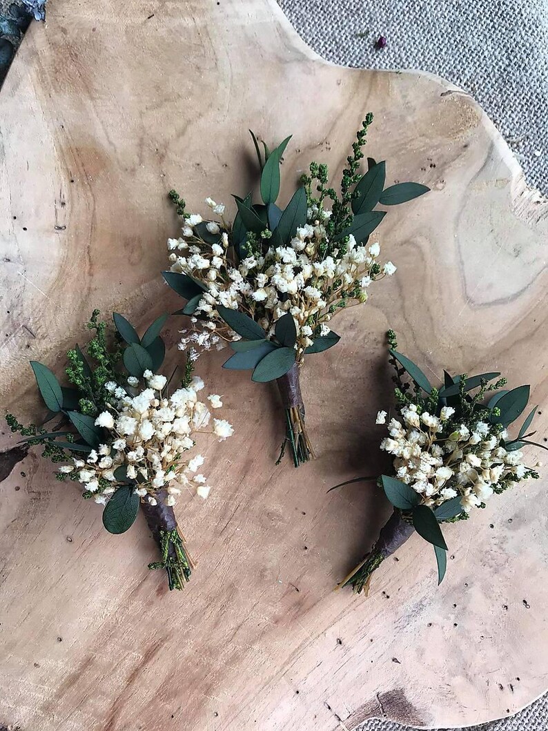 May include: Three small boutonnieres, each with tiny white flowers, green sprigs, and dark green leaves, bound with brown twine. They are displayed on a light brown wooden surface, ideal for weddings or special events.