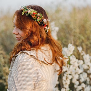 May include: A woman with long, red hair wearing a floral crown and a beige gingham shirt with a ruffled collar. She is standing in a field of white flowers.