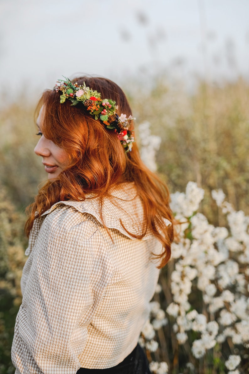 May include: A woman with long, red hair wearing a floral crown and a beige gingham shirt with a ruffled collar. She is standing in a field of white flowers.