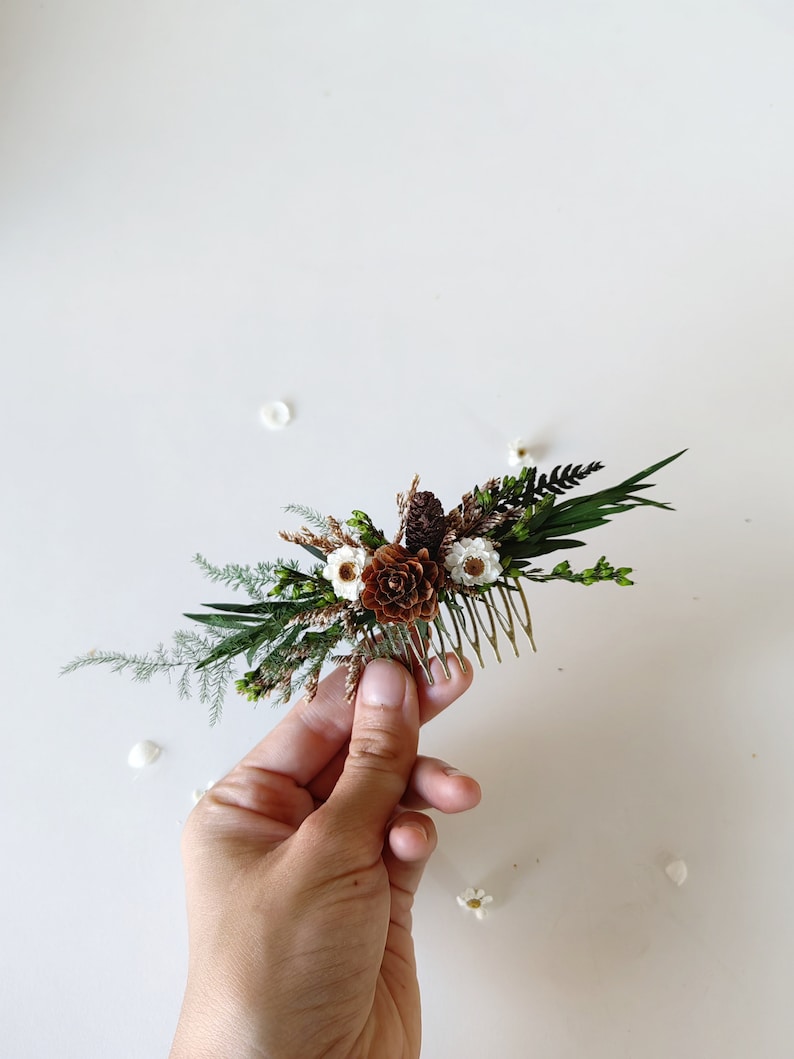 May include: A small floral hair comb with white flowers, brown pinecones, and green foliage. The comb is held in a hand against a white background.