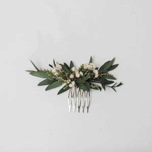 May include: A silver hair comb decorated with a floral arrangement. The arrangement includes dark green leaves and tiny white flowers. The comb is suitable for bridal or special occasion hairstyles.