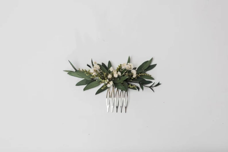 May include: A silver hair comb decorated with a floral arrangement. The arrangement includes dark green leaves and tiny white flowers. The comb is suitable for bridal or special occasion hairstyles.