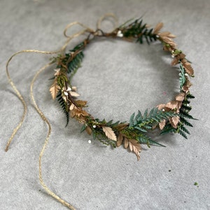 May include: A handcrafted floral crown featuring green and brown leaves, small white berries, and a twine tie. The crown is designed with a natural aesthetic, perfect for weddings or special occasions.