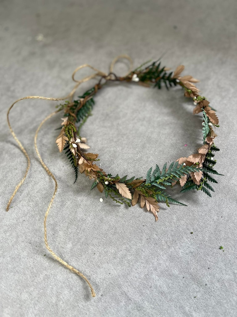 May include: A handcrafted floral crown featuring green and brown leaves, small white berries, and a twine tie. The crown is designed with a natural aesthetic, perfect for weddings or special occasions.