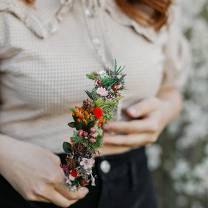 May include: A floral headband with a variety of dried flowers, including red berries, green moss, and small white flowers. The headband is held in the hand of a person wearing a beige shirt and black pants.