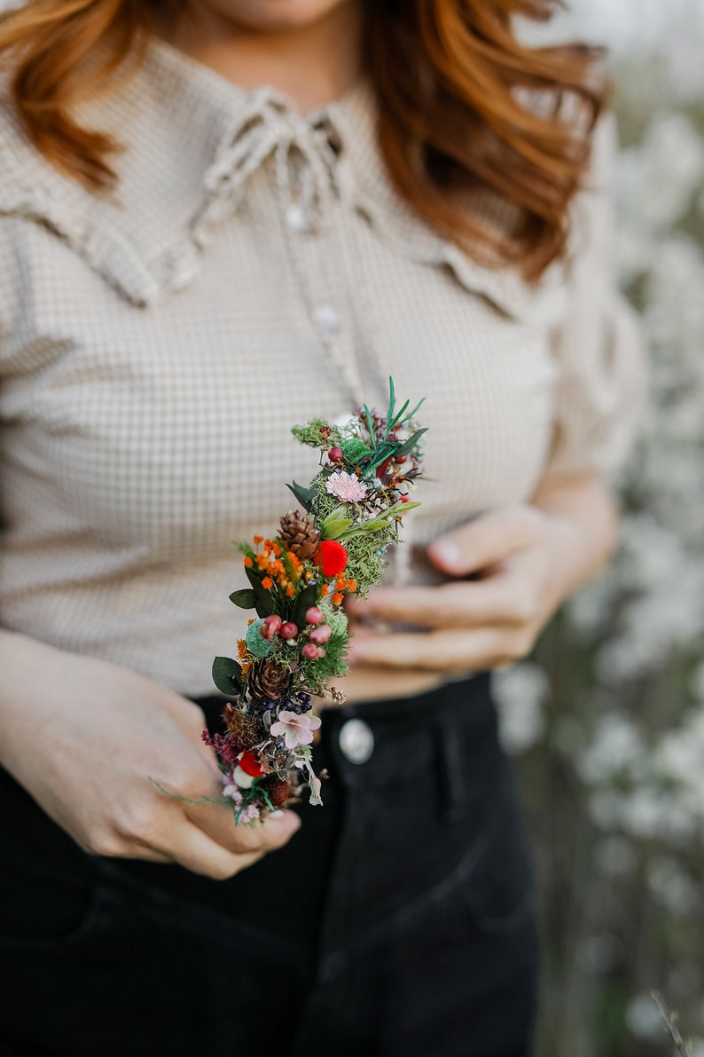 May include: A floral headband with a variety of dried flowers, including red berries, green moss, and small white flowers. The headband is held in the hand of a person wearing a beige shirt and black pants.
