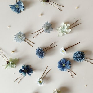 K&ouml;nnte beinhalten: Eine Auswahl an floralen Haarnadeln in verschiedenen Blau-, Wei&szlig;- und Gr&uuml;nt&ouml;nen. Jede Nadel hat ein anderes Blumendesign mit zarten Bl&uuml;tenbl&auml;ttern und einer braunen Metallnadel. Die Nadeln sind auf einer wei&szlig;en Oberfl&auml;che angeordnet.
