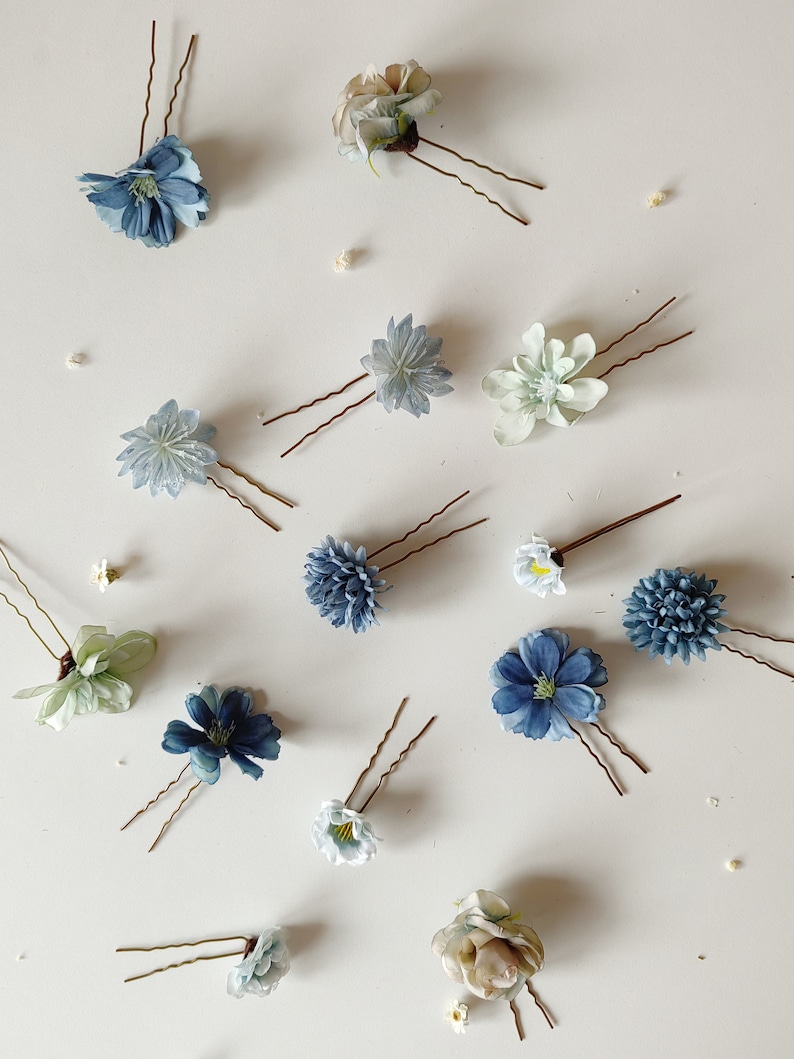 K&ouml;nnte beinhalten: Eine Auswahl an floralen Haarnadeln in verschiedenen Blau-, Wei&szlig;- und Gr&uuml;nt&ouml;nen. Jede Nadel hat ein anderes Blumendesign mit zarten Bl&uuml;tenbl&auml;ttern und einer braunen Metallnadel. Die Nadeln sind auf einer wei&szlig;en Oberfl&auml;che angeordnet.