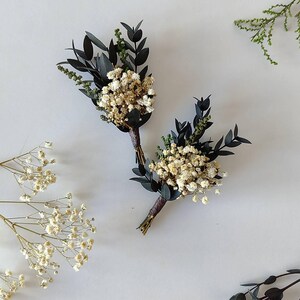 May include: Two small boutonnieres, each featuring dried white flowers, dark green leaves, and dark brown stems. They are displayed on a white surface, with additional sprigs of greenery. Ideal for weddings or special occasions.