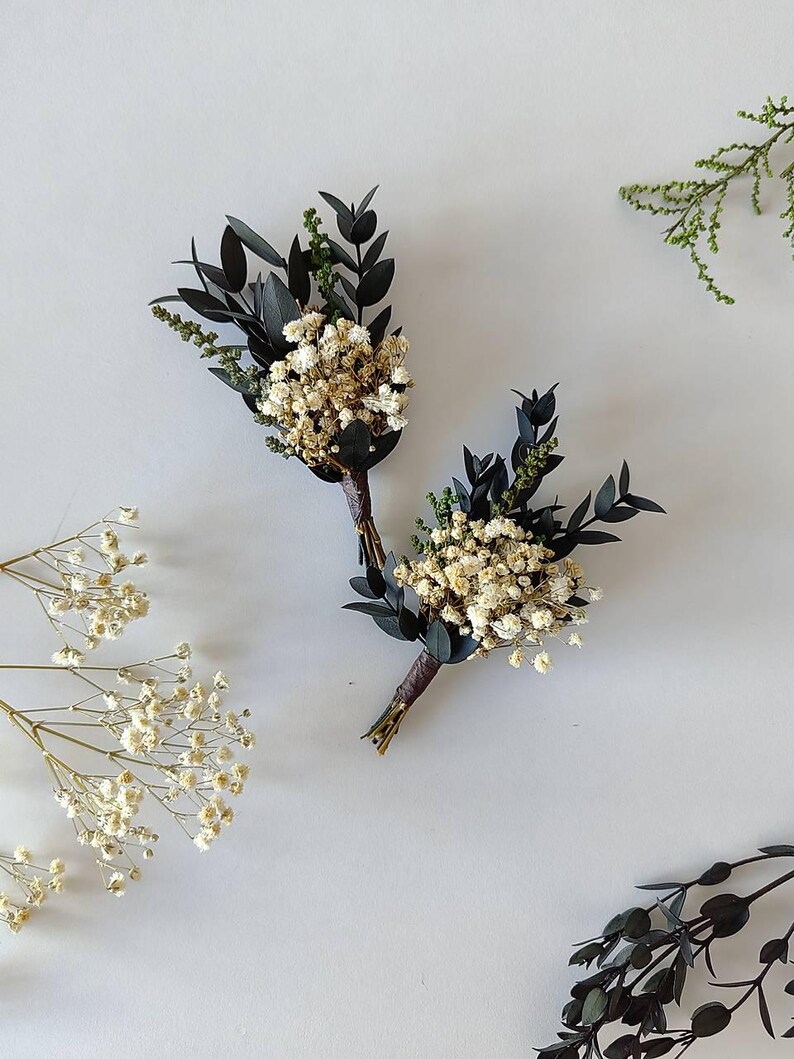 May include: Two small boutonnieres, each featuring dried white flowers, dark green leaves, and dark brown stems. They are displayed on a white surface, with additional sprigs of greenery. Ideal for weddings or special occasions.