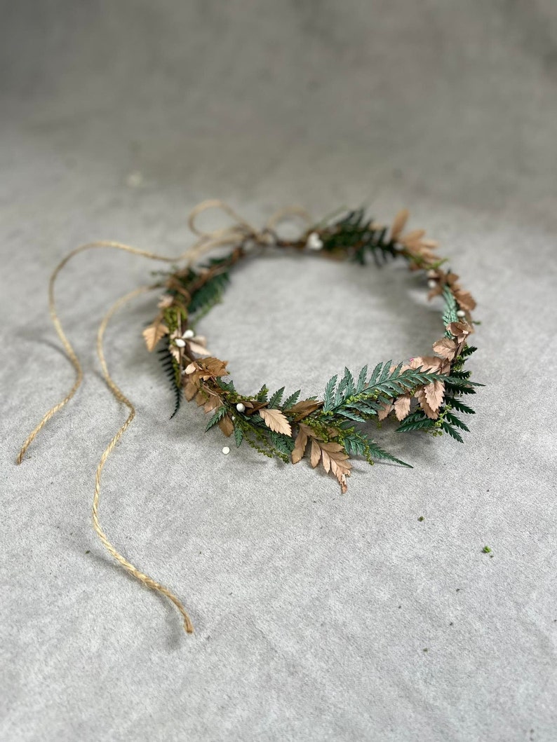 May include: A handcrafted floral crown featuring a circular design. The crown is adorned with green fern leaves, gold-toned leaves, and small white bead accents. Natural twine is used to tie the crown together.