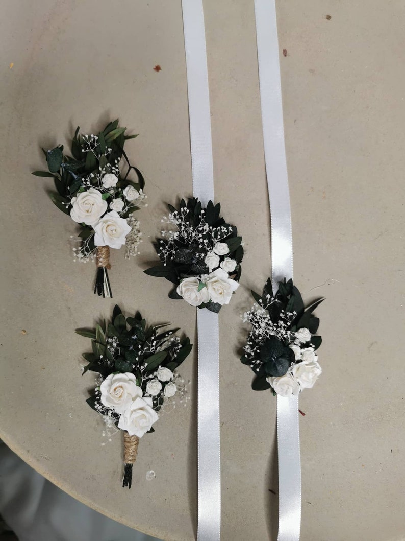 Puede incluir: Cuatro arreglos florales. Dos son ramos de rosas blancas, hojas verdes y paniculata, envueltos en hilo. Dos son ramilletes de mu&ntilde;eca con cintas de raso blancas. Las flores est&aacute;n sobre un fondo neutro.