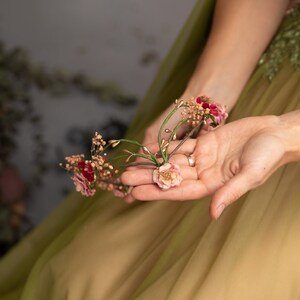 May include: A hand holding a small bouquet of pink and red flowers with gold accents. The flowers are arranged on a green stem.