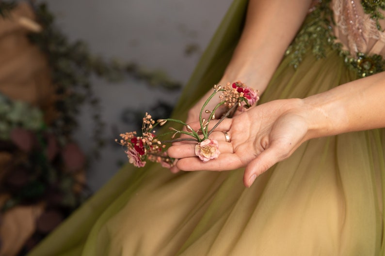 May include: A hand holding a small bouquet of pink and red flowers with gold accents. The flowers are arranged on a green stem.