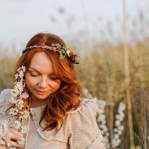 May include: A woman with long, red hair wears a floral crown and a light brown, checkered dress. She is holding a sprig of white flowers in her hand and looking down at them.